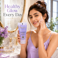 A woman in a lavender satin robe applies face wash cream to her cheek while holding a tube of Advanced Face Wash by The Skincare Institute. She is standing in a bright, modern bathroom with gold fixtures, a mirror, and a vase of purple flowers.